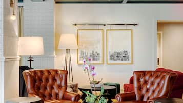 A modern hotel lobby with leather armchairs, a round table with a vase of flowers, and framed cityscape pictures on the wall.