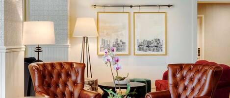 Un moderno vestíbulo de hotel con sillones de cuero, una mesa redonda con un florero y fotografías enmarcadas de la ciudad en la pared.