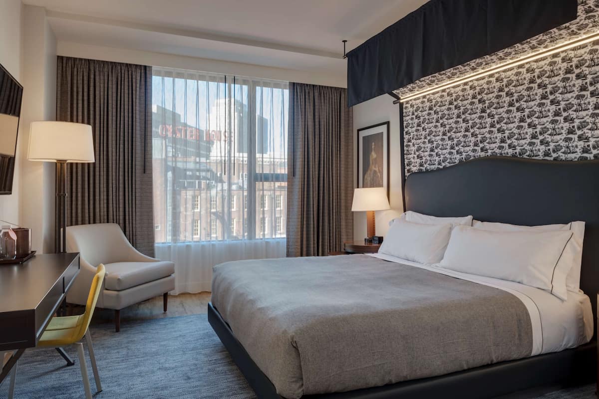 A hotel room with a large bed, a desk with a chair, a window with curtains, and a patterned wall.