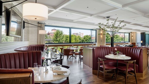 A restaurant with booth seating, round tables, and a view of the city through large windows. - Canopy By Hilton Boston Downtown (Boston)