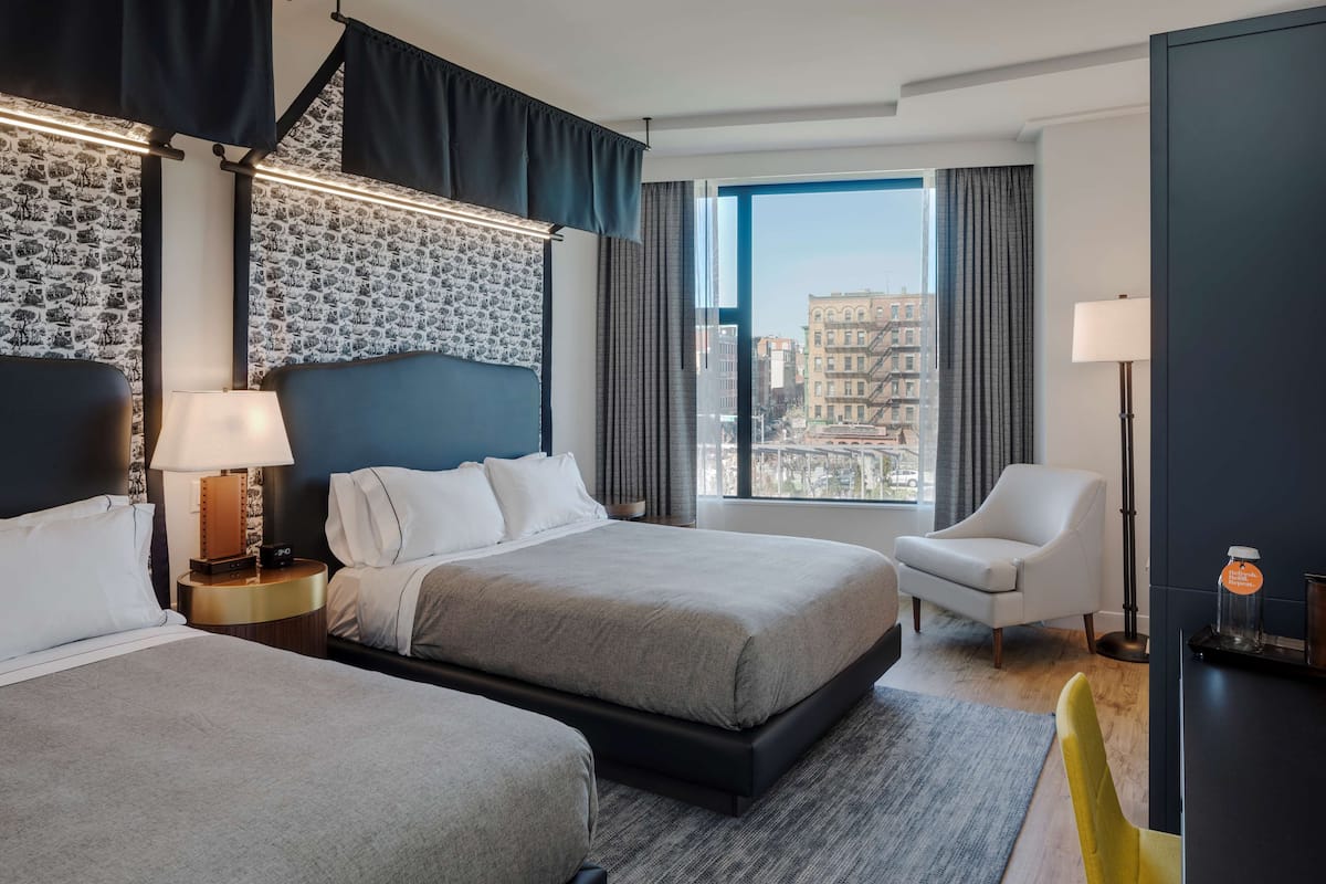 A hotel room with two beds, a view of the city, a modern lamp, and a chair.