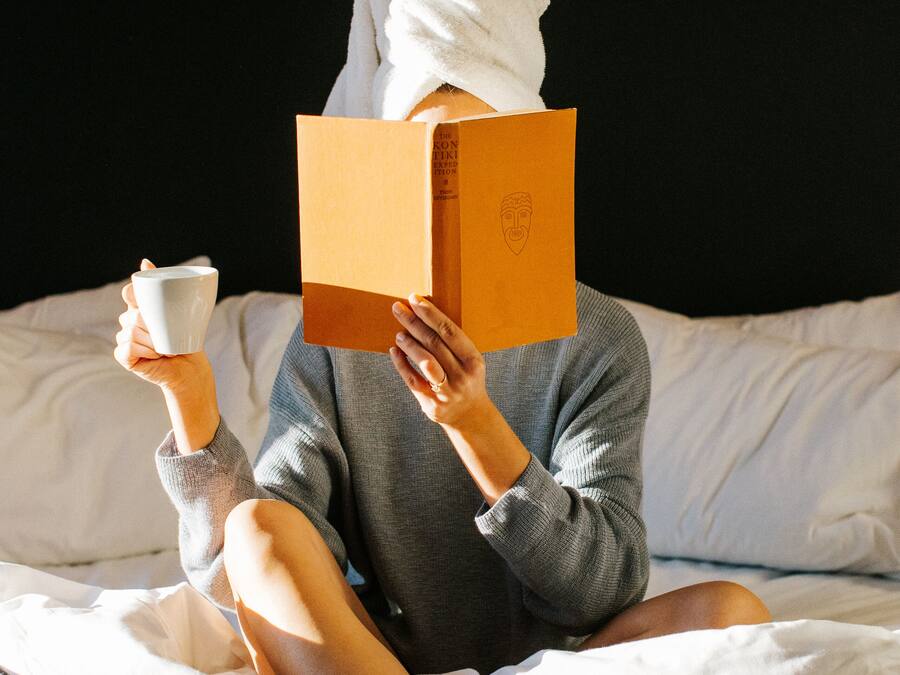 A person sitting on a bed with a book and a cup, wearing striped socks and a towel on their head.