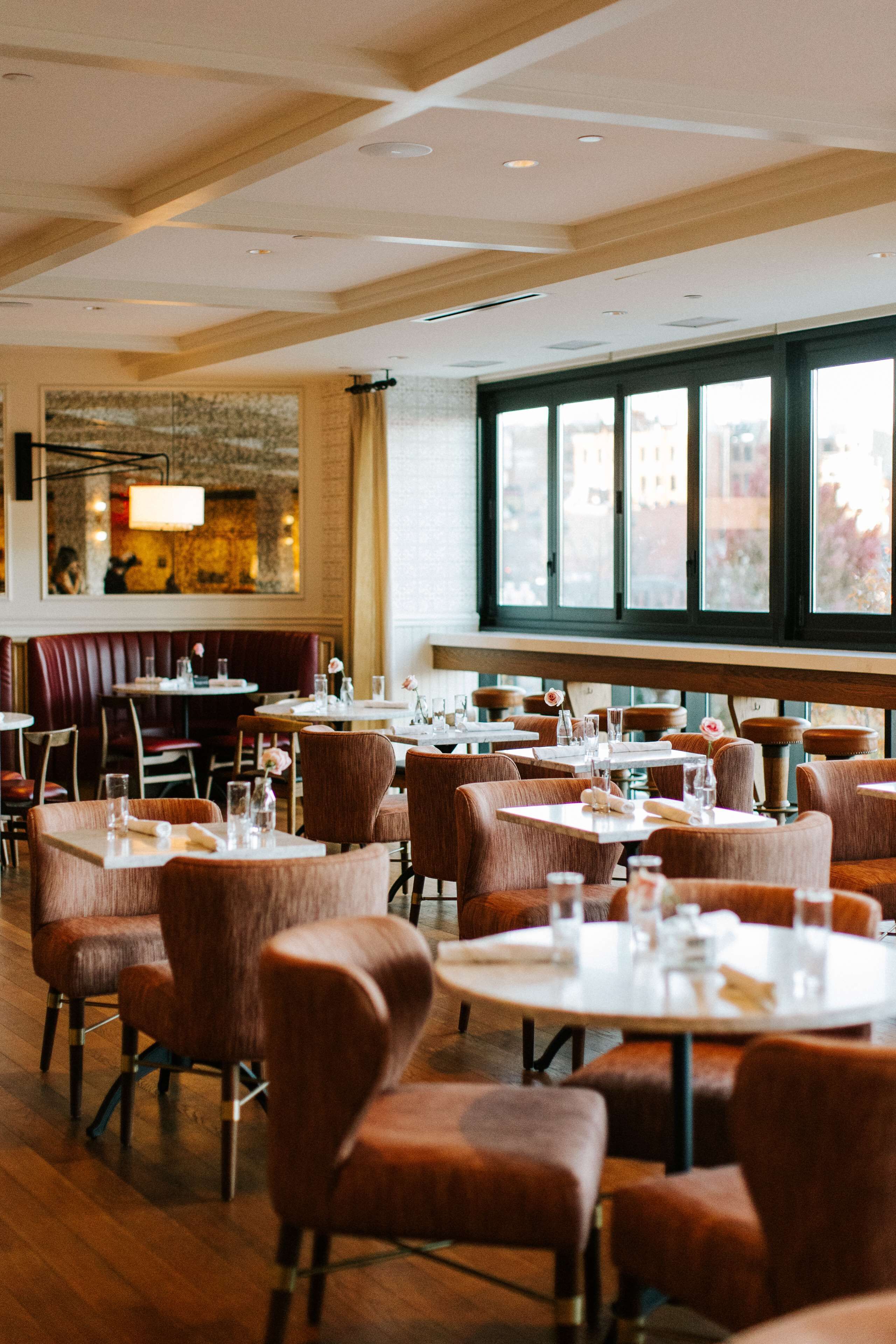 Un restaurant avec des tables rondes et des banquettes, des planchers en bois et de grandes fenêtres offrant une vue sur la ville.