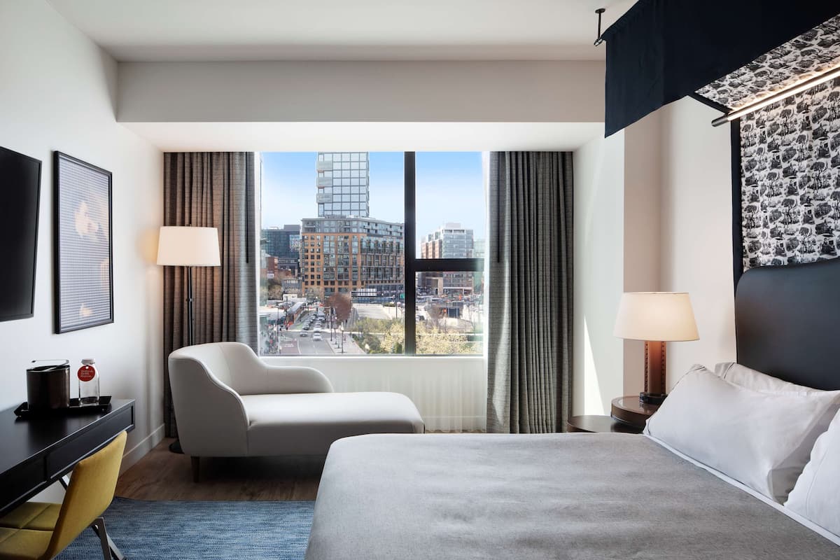 A hotel room with a large bed, a desk, a chair, and a view of the cityscape through large windows.