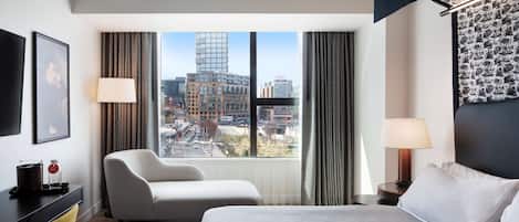 A hotel room with a large bed, a desk, a chair, and a view of the cityscape through large windows.