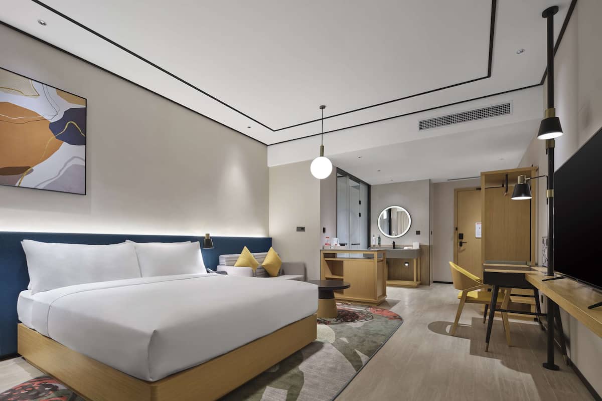 A modern hotel room with a large bed, a desk, a chair, a mirror, and a TV.