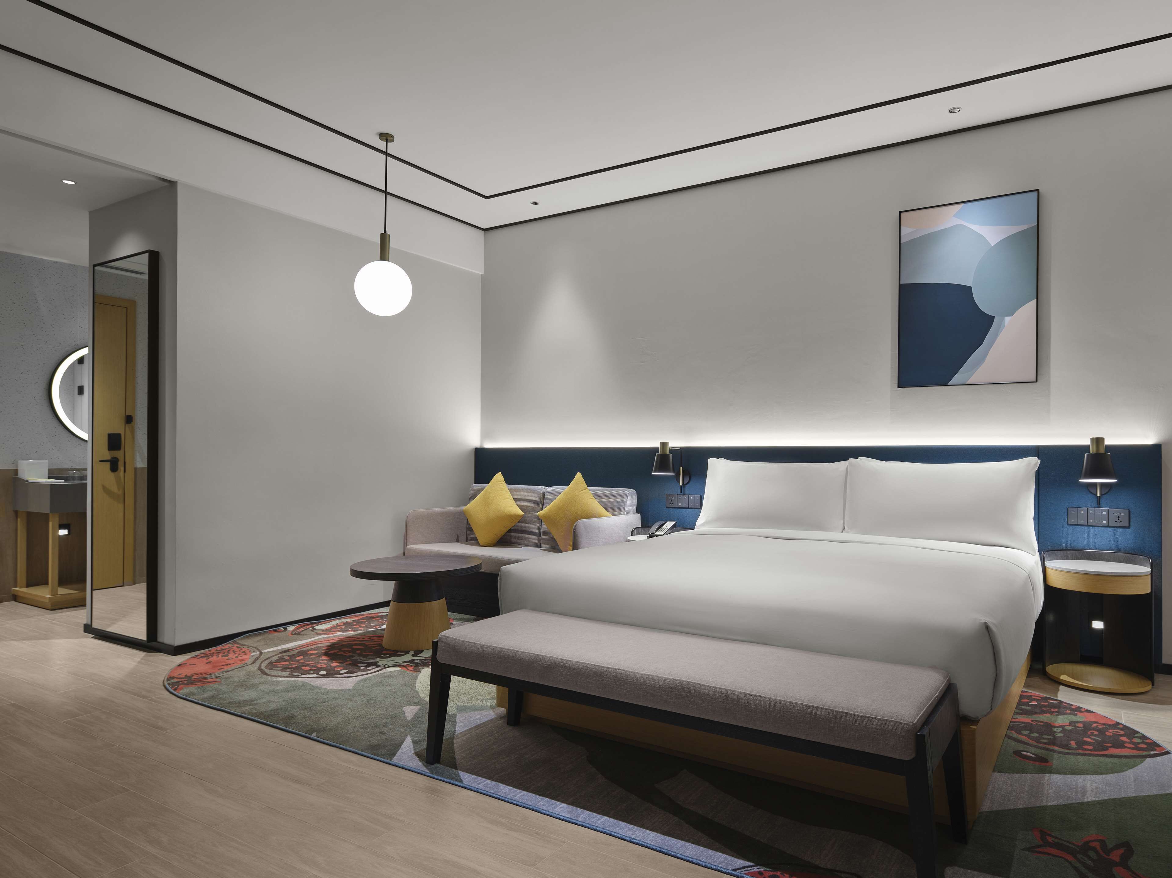 Een moderne hotelkamer met een groot bed, nachtkastjes, een bankje, een sofa, een ronde tafel, een spiegel en een kunstwerk aan de muur.