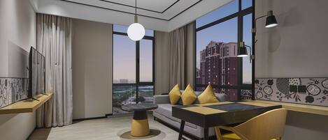 A modern hotel room with a large window, a dining area with yellow chairs, a round table, and a sofa with yellow cushions.