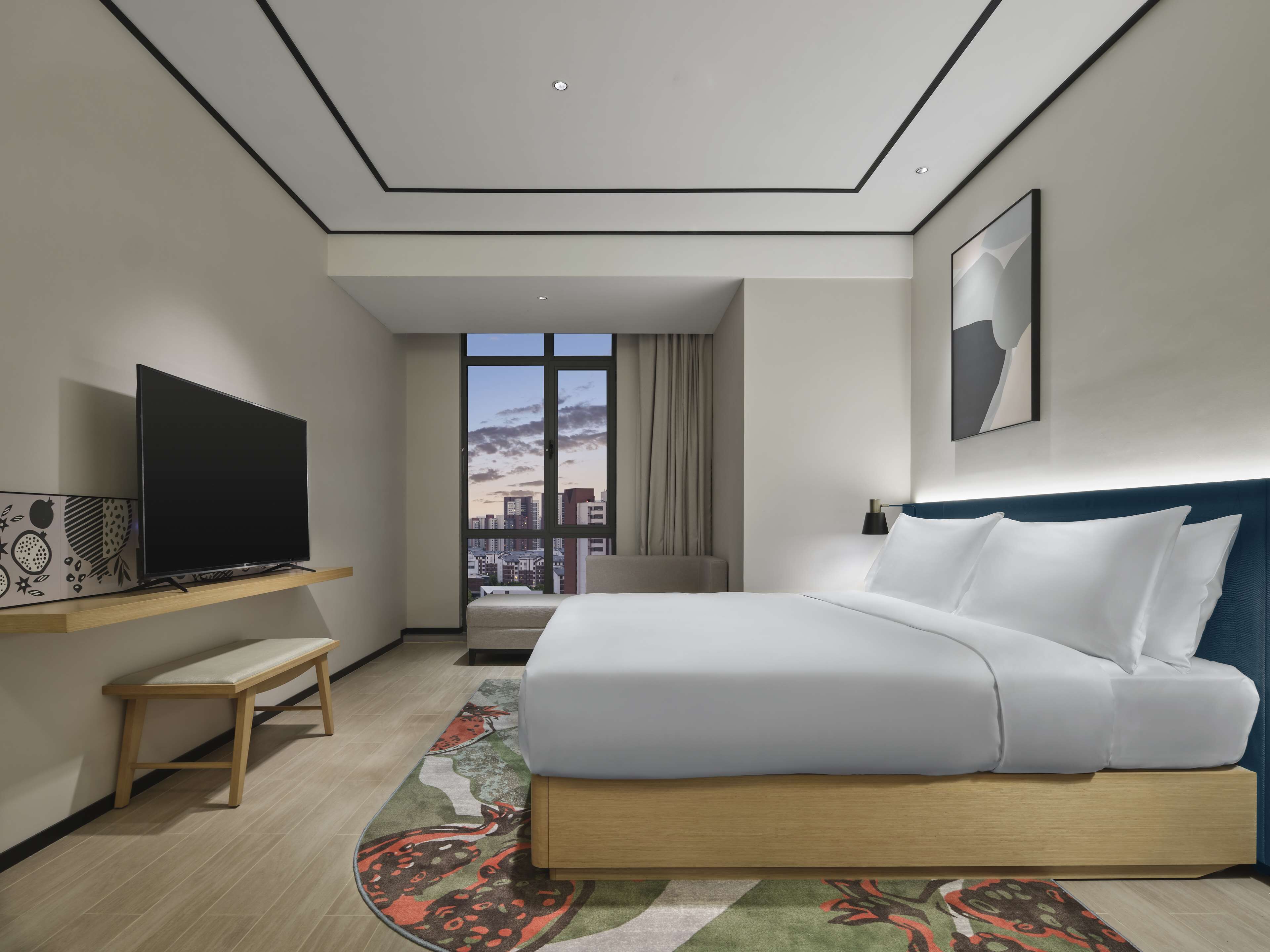 A modern hotel room with a large bed, a flat-screen TV on a wooden shelf, a city view through a large window, and a blue headboard.