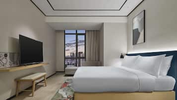 A modern hotel room with a large bed, a flat-screen TV on a wooden shelf, a city view through a large window, and a blue headboard.