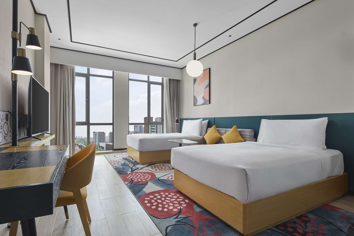 A modern hotel room with a large bed, a desk, and a chair. There is a view of the city through large windows.