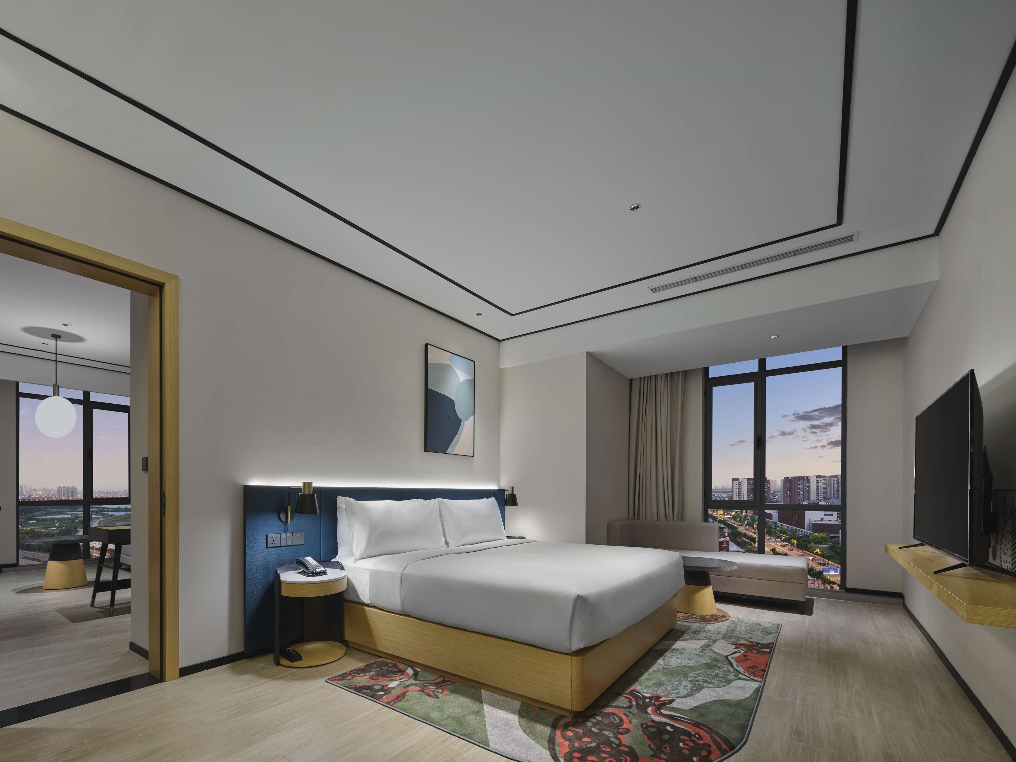 Habitación de hotel moderna con una cama grande, un escritorio, una silla, televisión y vistas a la ciudad a través de amplios ventanales.