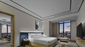 A modern hotel room with a large bed, a desk, a chair, a TV, and a view of the city through large windows.