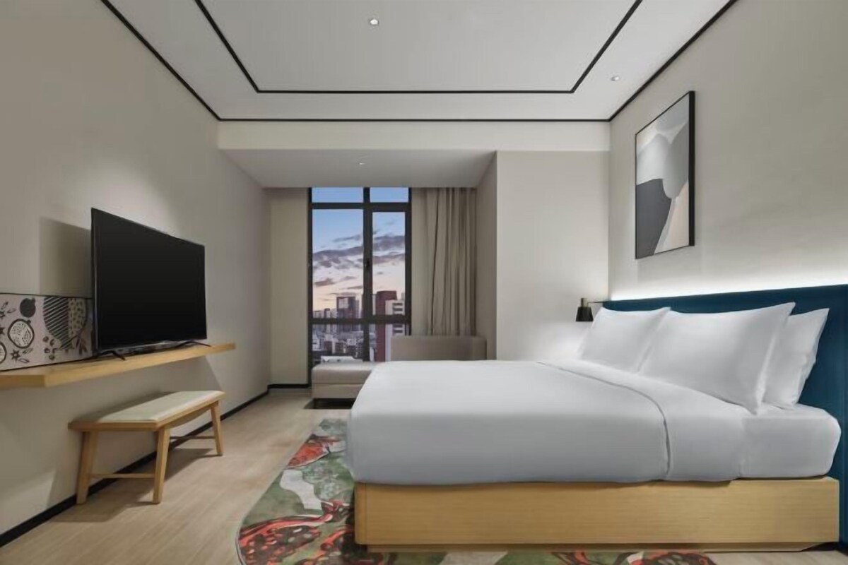 Een moderne hotelkamer met een groot bed, een televisie op een plank, een raam met uitzicht op de stad en een plafond met een geometrisch patroon.