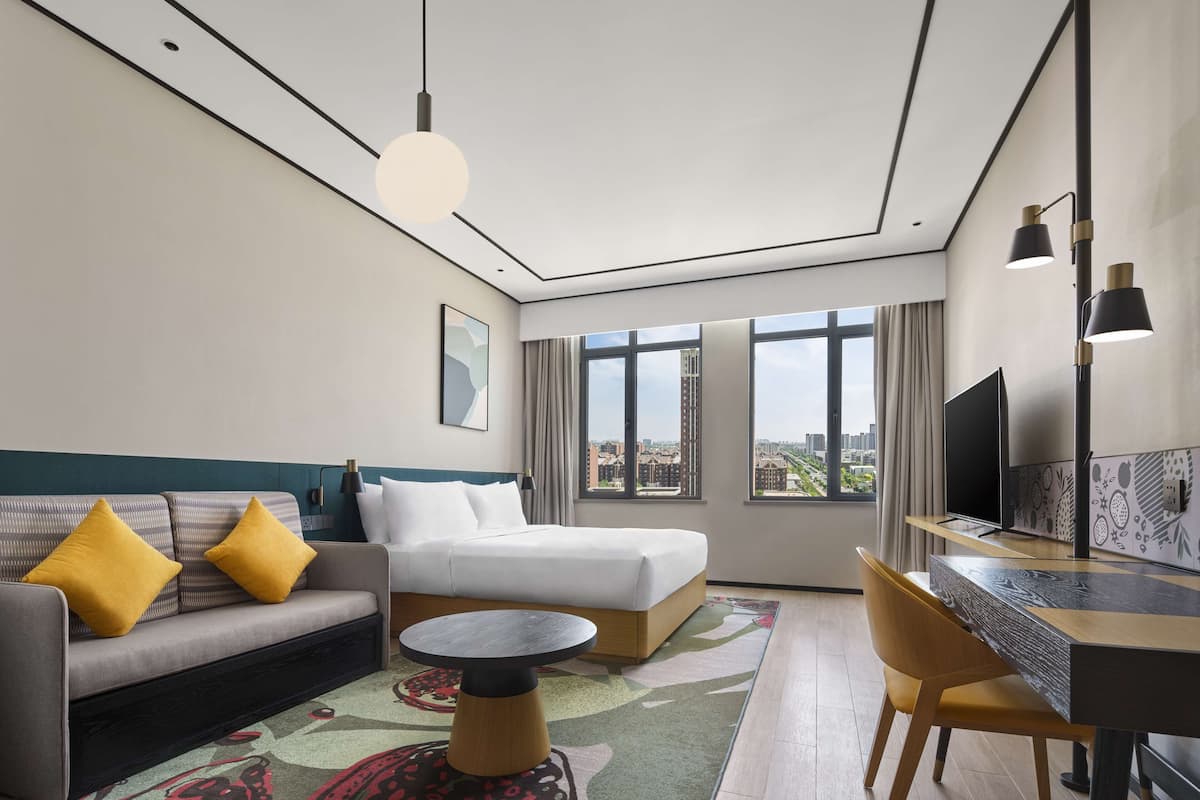 A modern hotel room with a bed, sofa, desk, and large windows offering city views.