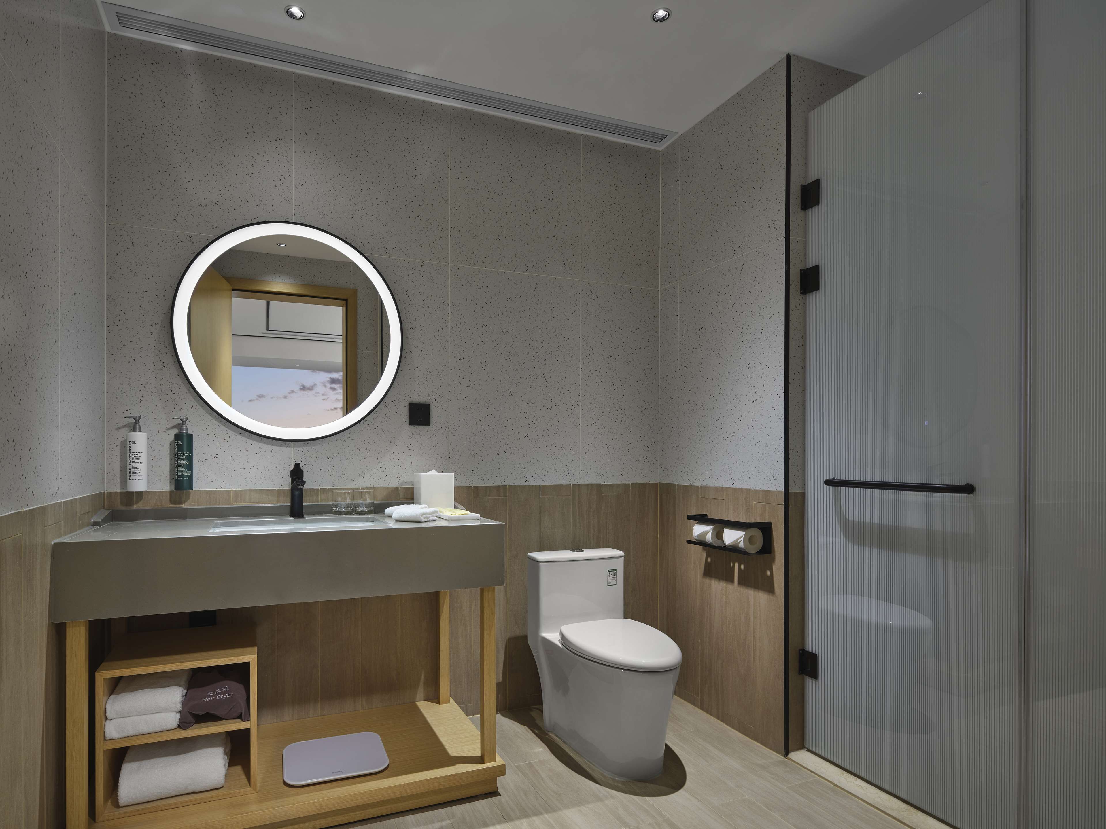 Un baño moderno con tocador de madera, un espejo redondo, inodoro y ducha a ras de suelo.