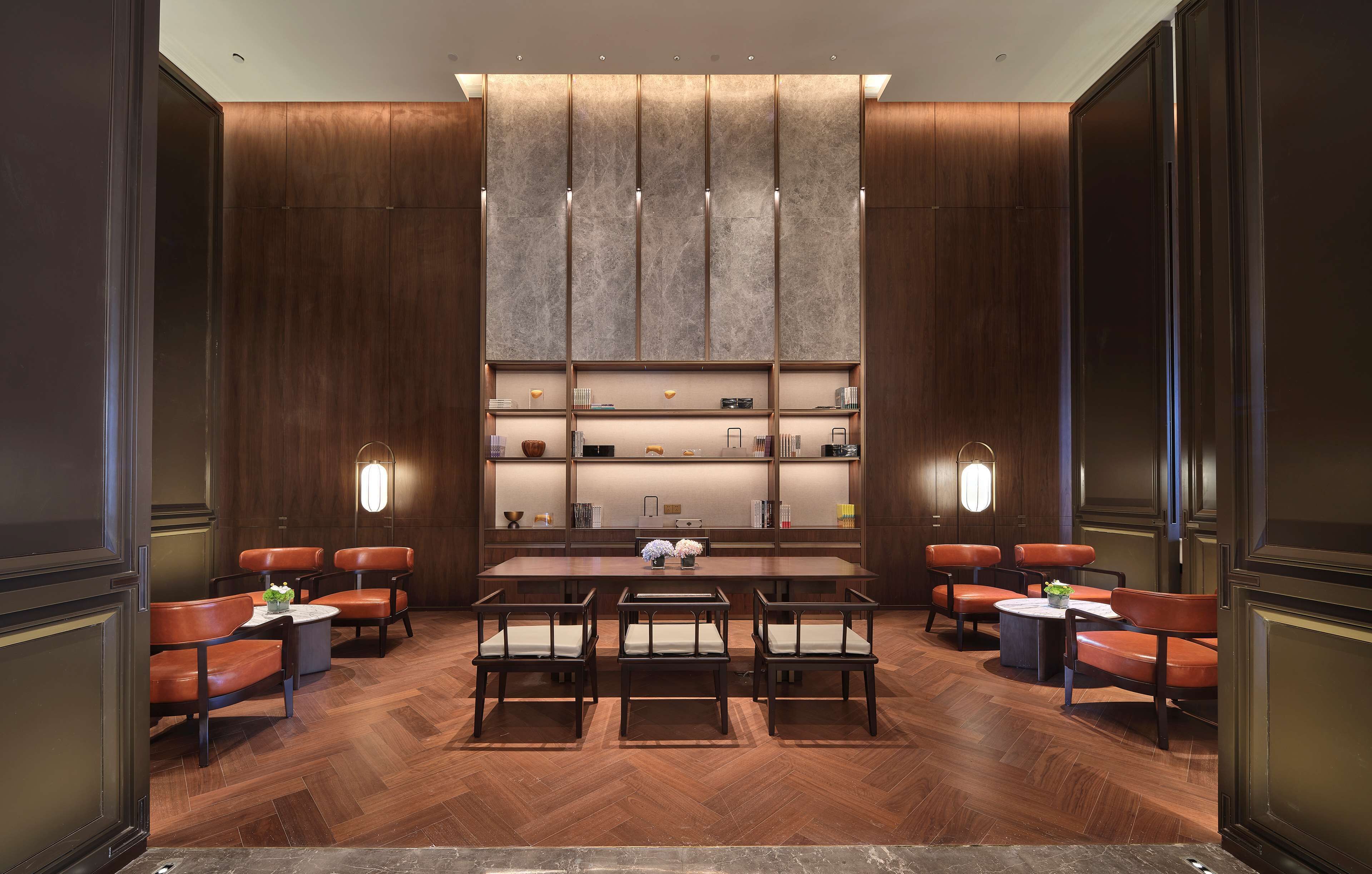 A modern, well-lit lounge area with a long table and multiple chairs, surrounded by wooden paneling and shelves with various items.