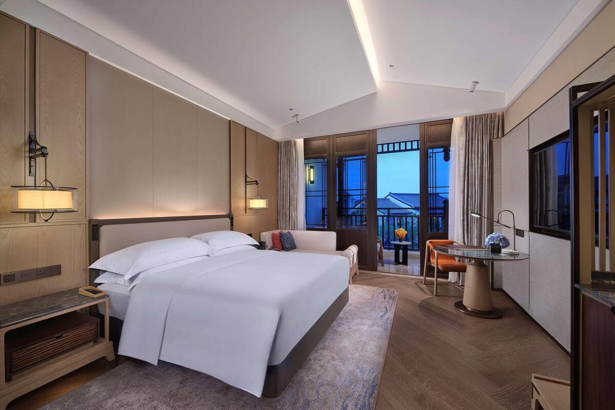 Camera d'albergo con un grande letto, comodini, una scrivania con una sedia, un balcone con vista e un'ampia finestra.