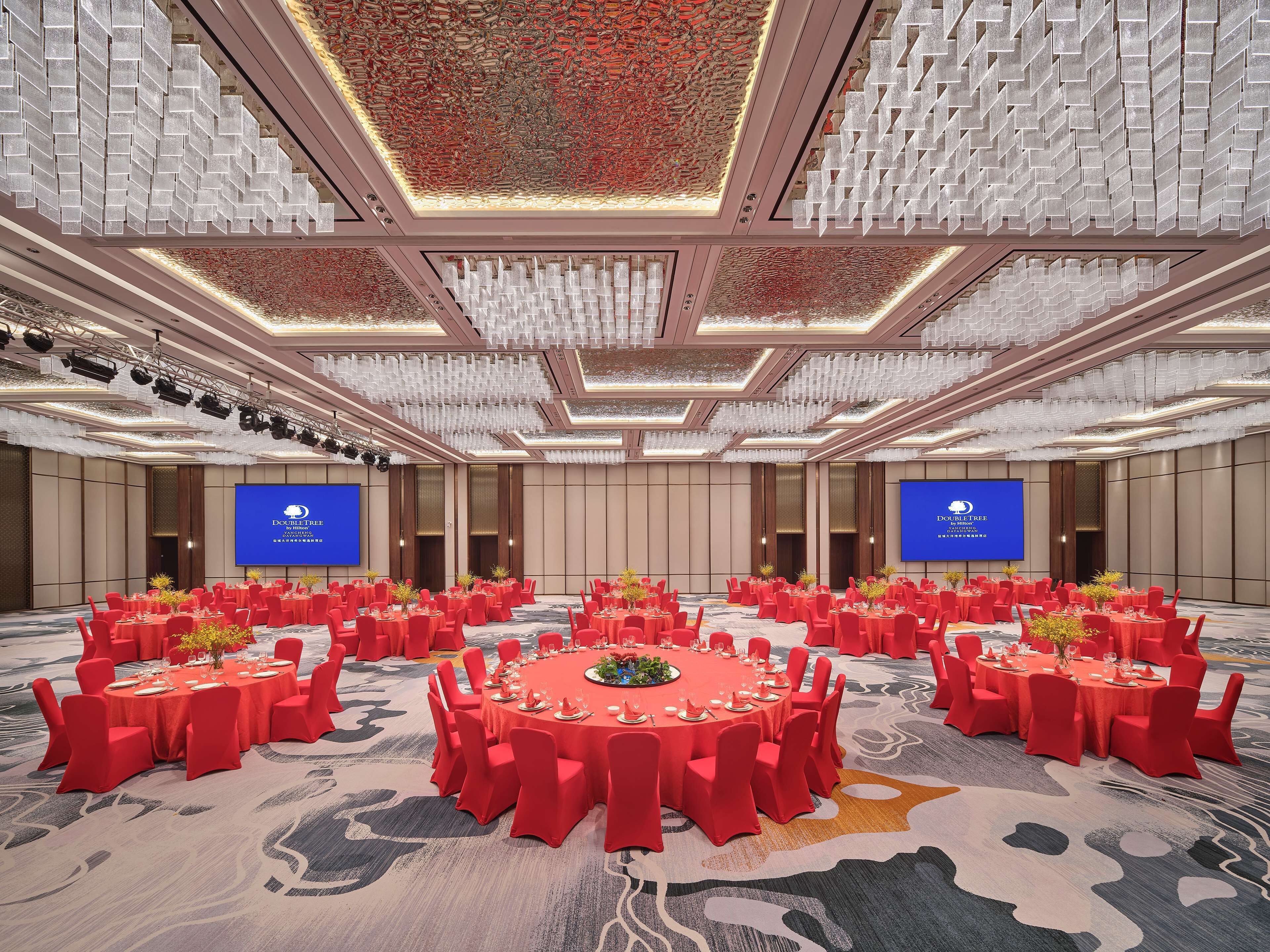 A conference room with multiple round tables covered in red tablecloths, equipped with chairs and centerpieces.