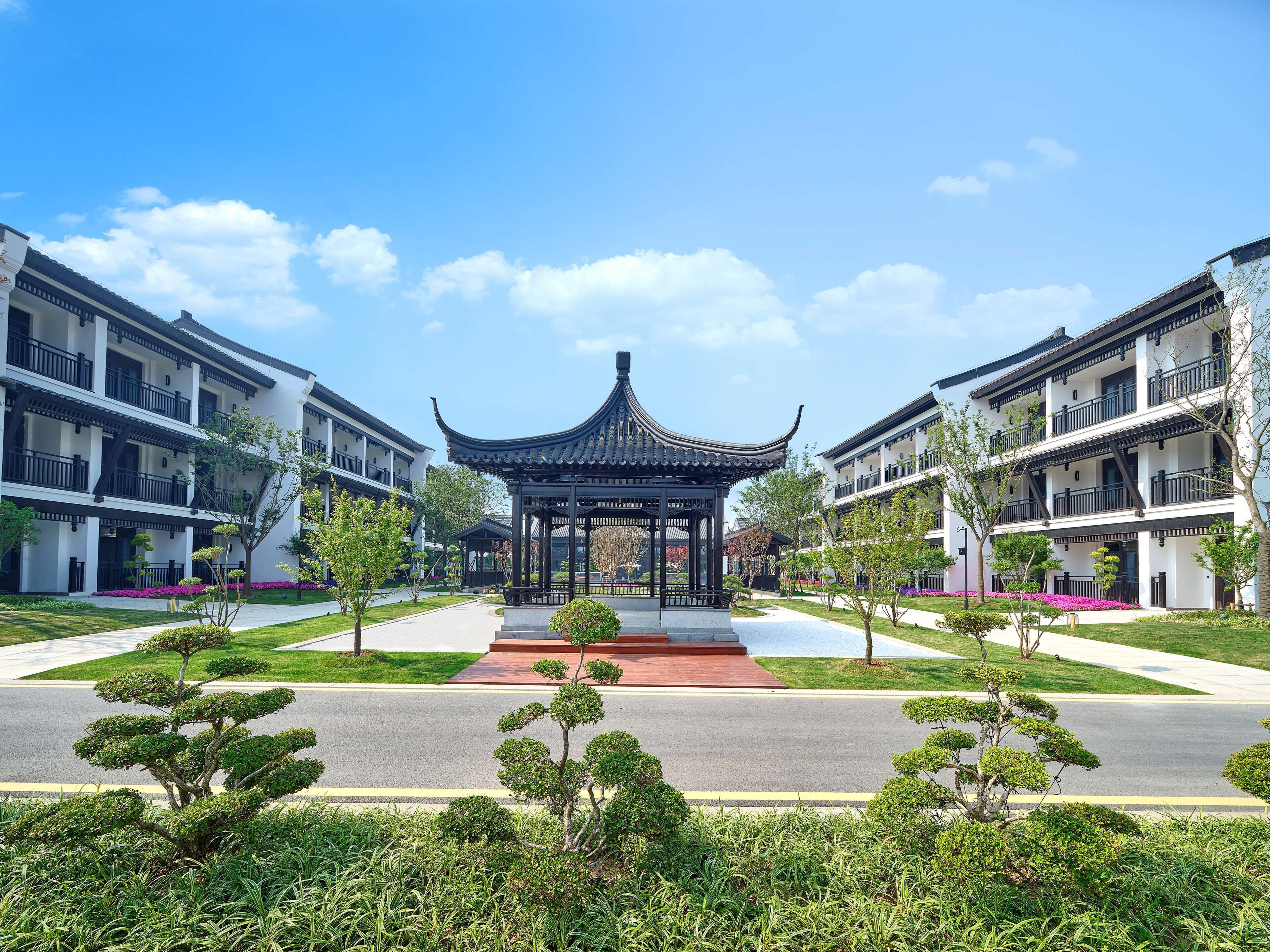 A traditional Chinese pavilion with a red base, surrounded by modern buildings with balconies and a well-maintained garden with various plants.