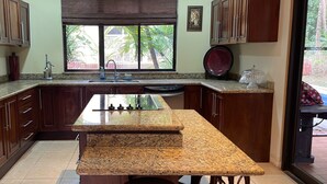 A kitchen with granite countertops, wooden cabinets, a stainless steel refrigerator, a window with a view of palm trees, and a red plate on the counter. - Casa Safari Maganda located in the middle of
paradise. (Tarcoles)