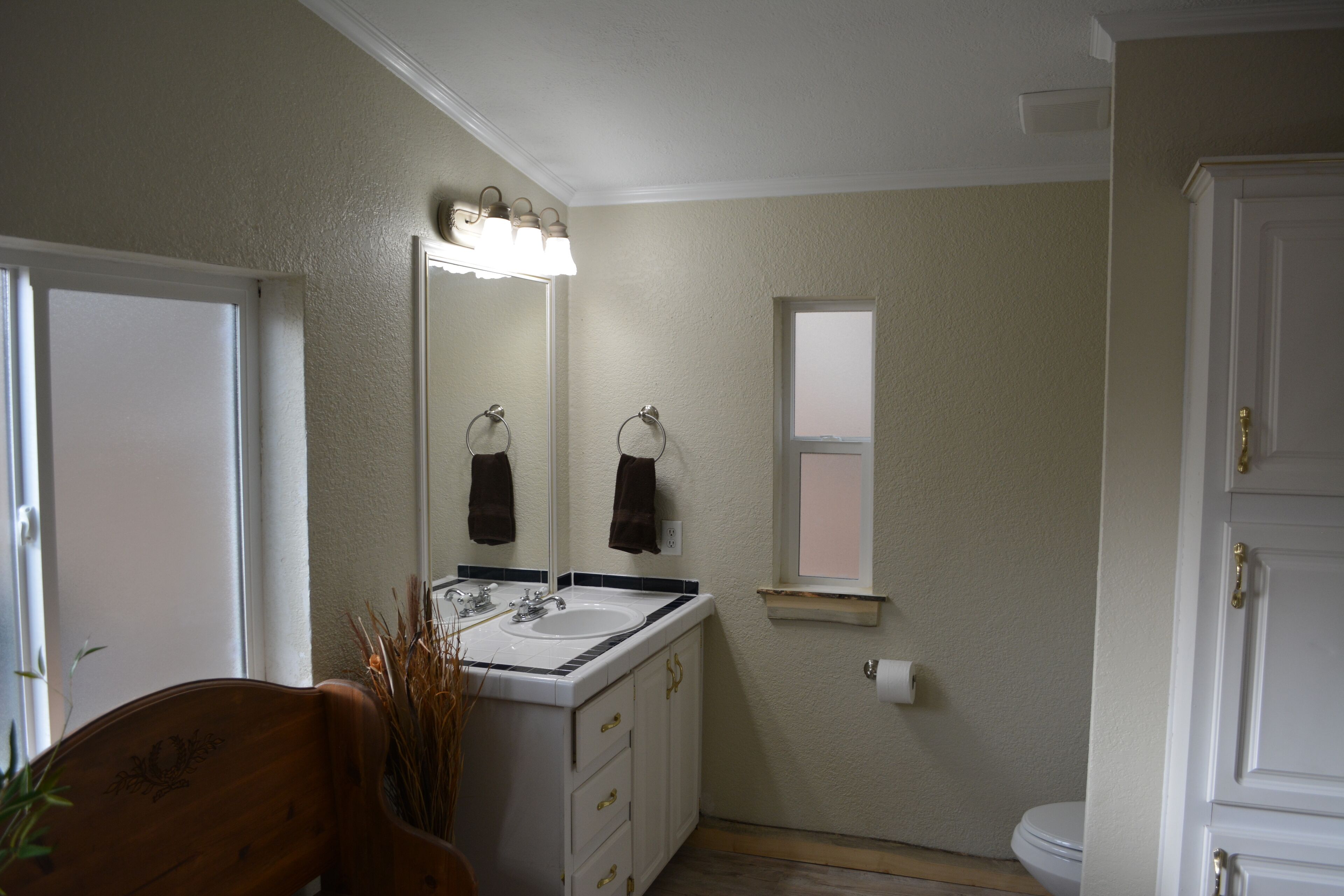 A bathroom with a white sink, mirror, and a window. There is a wooden chair and a plant near the sink.