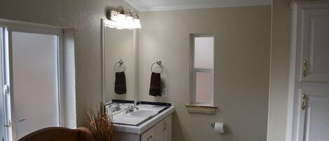 A bathroom with a white sink, mirror, and a window. There is a wooden chair and a plant near the sink.