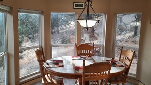 A dining area with a round table and six wooden chairs, large windows offering a view of a wooded area, and a hanging light fixture. - Forest cabin on 4 acres,  Newly renovated, Quiet and secluded. (Florissant)