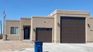 A residential building with a garage, a front door with a decorative glass panel, and a blue trash bin in the foreground. - Brand new home,  55 ft garage. 3 bedrooms, sleeps 10.  18ft high garage door.  (Lake havasu)