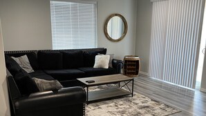 A modern living room with a black leather sofa, a coffee table, and a wooden side table. There is a round mirror on the wall, a ceiling fan, and a large window with blinds. - Brand new home,  55 ft garage. 3 bedrooms, sleeps 10.  18ft high garage door.  (Lake havasu)