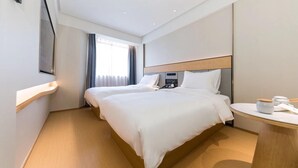 A hotel room with two beds, a bedside table, a TV mounted on the wall, and a window with curtains.