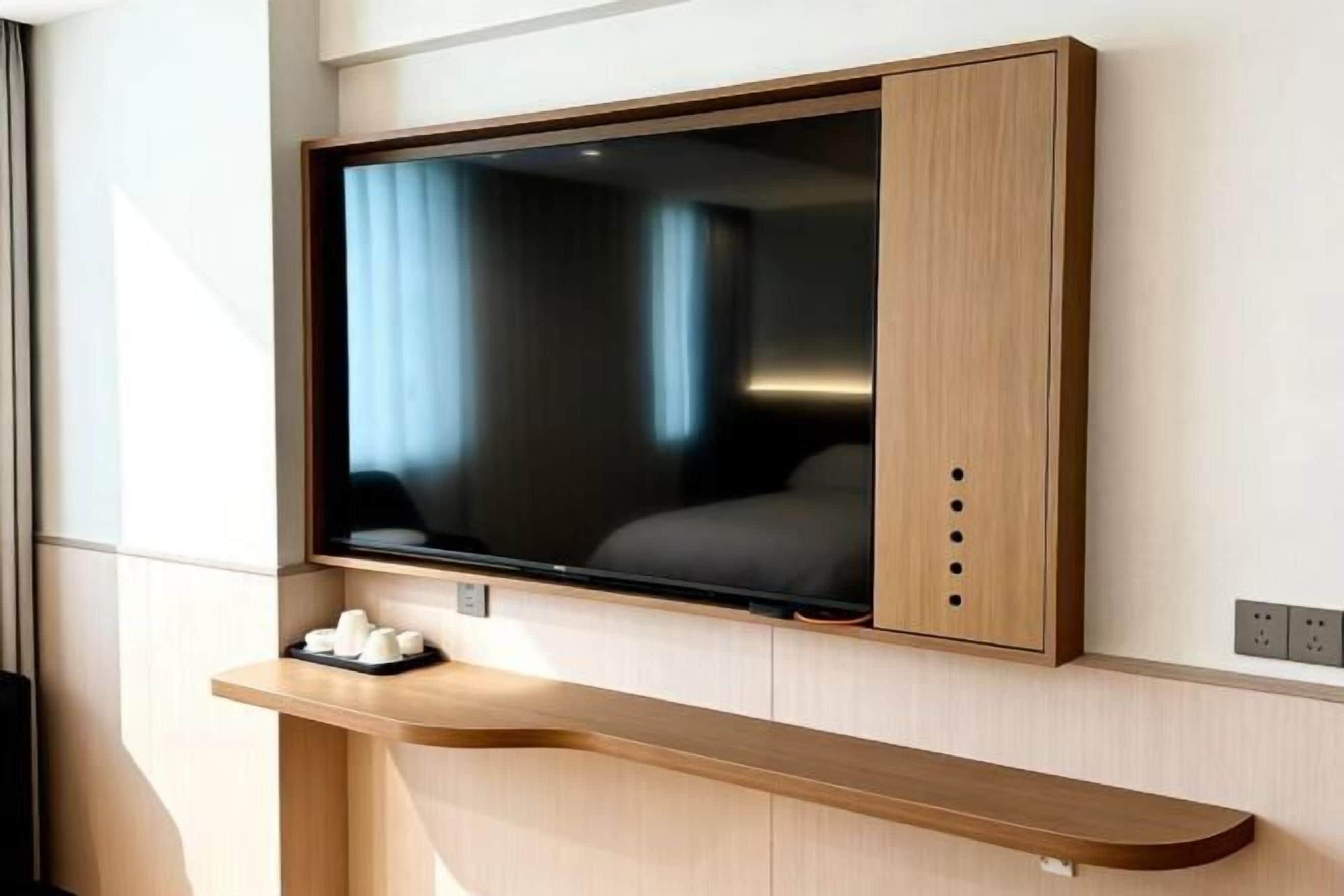 A modern hotel room with a large flat-screen TV mounted on a wooden wall, a wooden console table below, and a power outlet to the right.