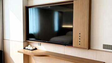 A modern hotel room with a large flat-screen TV mounted on a wooden wall, a wooden console table below, and a power outlet to the right.
