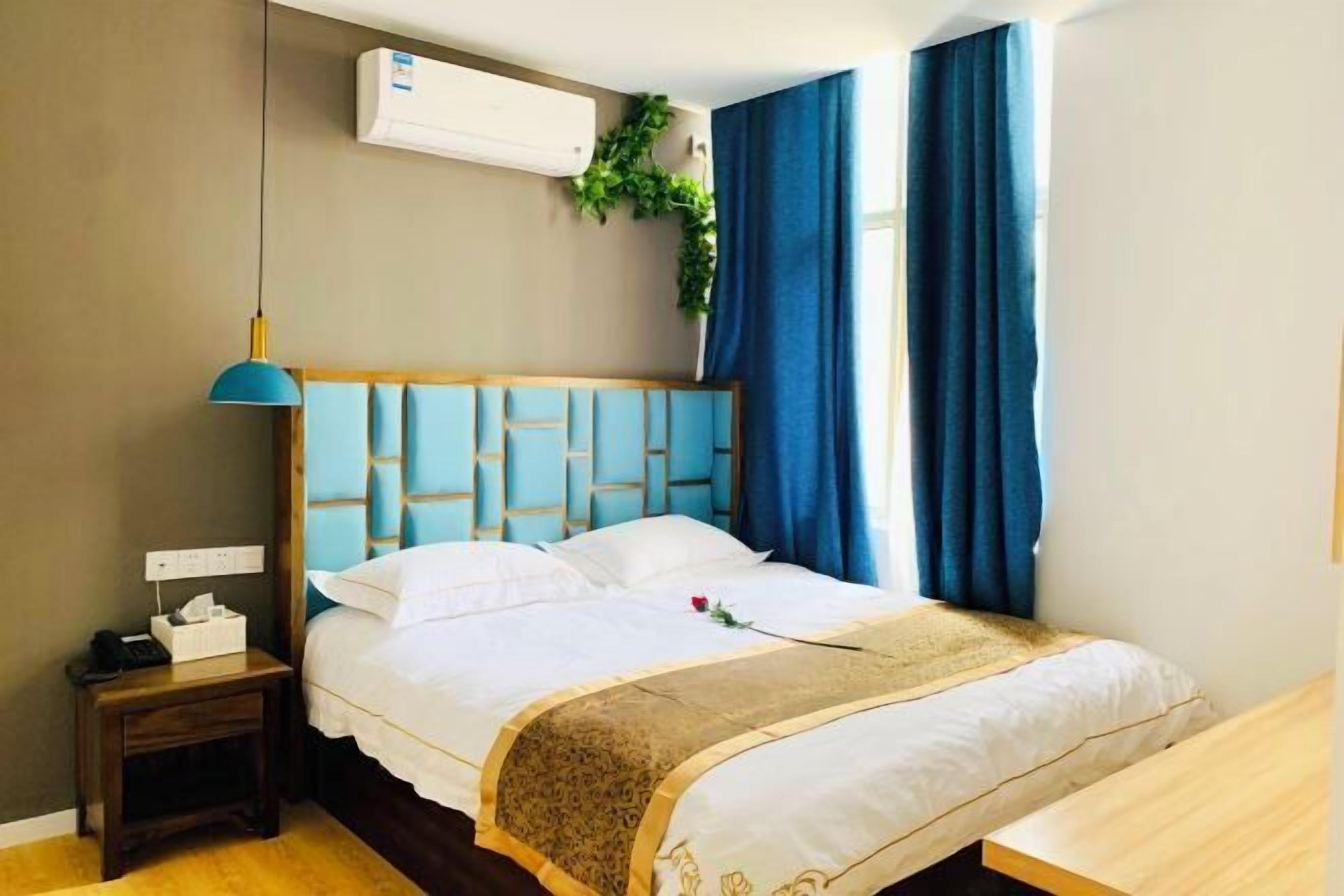 A hotel room with a bed, bedside table, lamp, air conditioning unit, and a window with curtains.