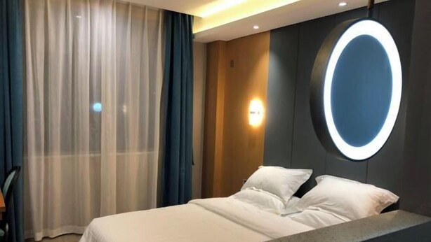 A hotel room with a bed, a nightstand, a wall-mounted light, and a window with sheer curtains. - Nanchang 520 Youth Hotel (Mailu Campus Center store of University of Finance and Economics) (Nanchang)