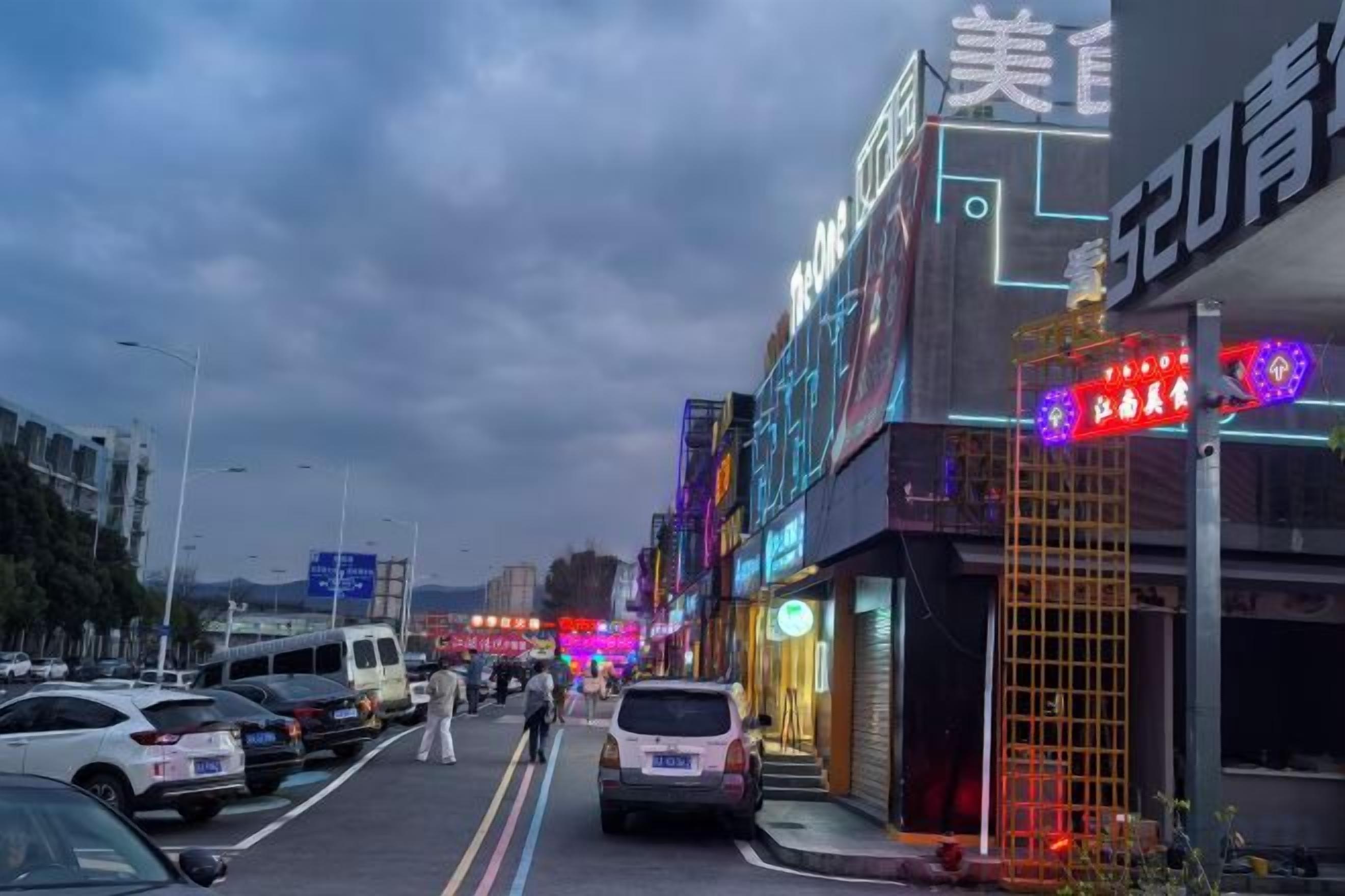 Una calle con coches aparcados, un peatón y escaparates iluminados.