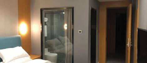 A modern hotel room with a bed, a glass shower enclosure, and a wooden door leading to another room.