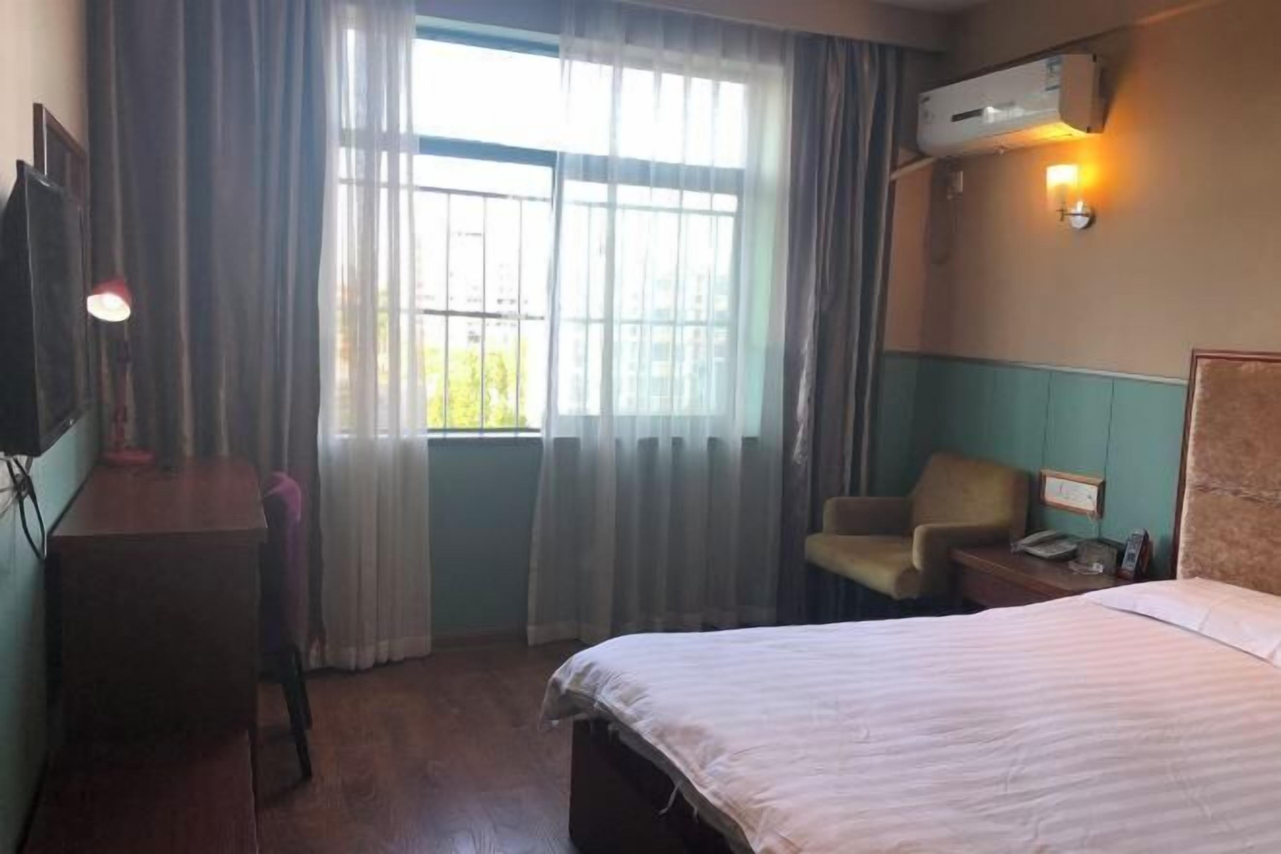 A hotel room with a bed, a desk, a chair, a window with curtains, and an air conditioning unit.
