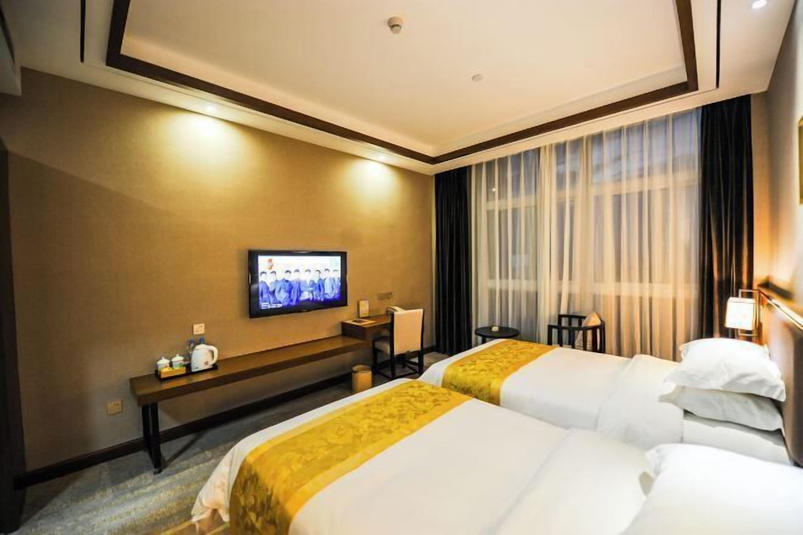 A hotel room with two beds, a television on the wall, a desk with a chair, and a window with curtains.