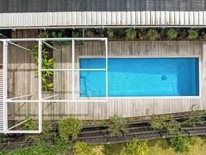 A rectangular swimming pool with a wooden deck surrounding it, set against a backdrop of a wooden fence and greenery.