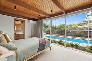 A bedroom with a large window, a bed, a bedside table, and a view of a swimming pool.