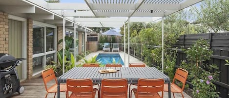 A covered outdoor patio with a pool, surrounded by a wooden deck and orange chairs.