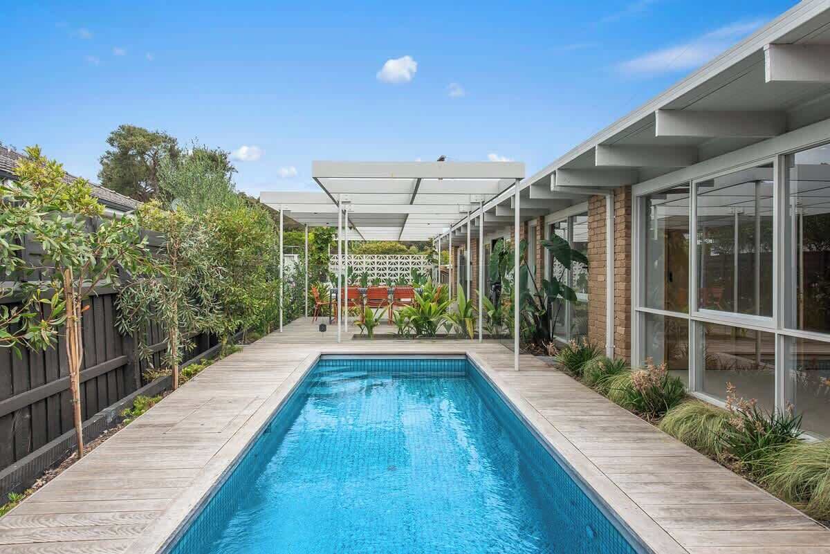 A rectangular swimming pool with a wooden deck, surrounded by a wooden fence and plants.