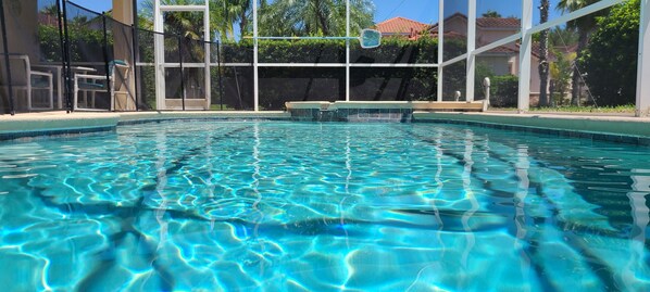 A clear swimming pool with a view of the surrounding area, including a covered seating area and a building with a red-tiled roof.
