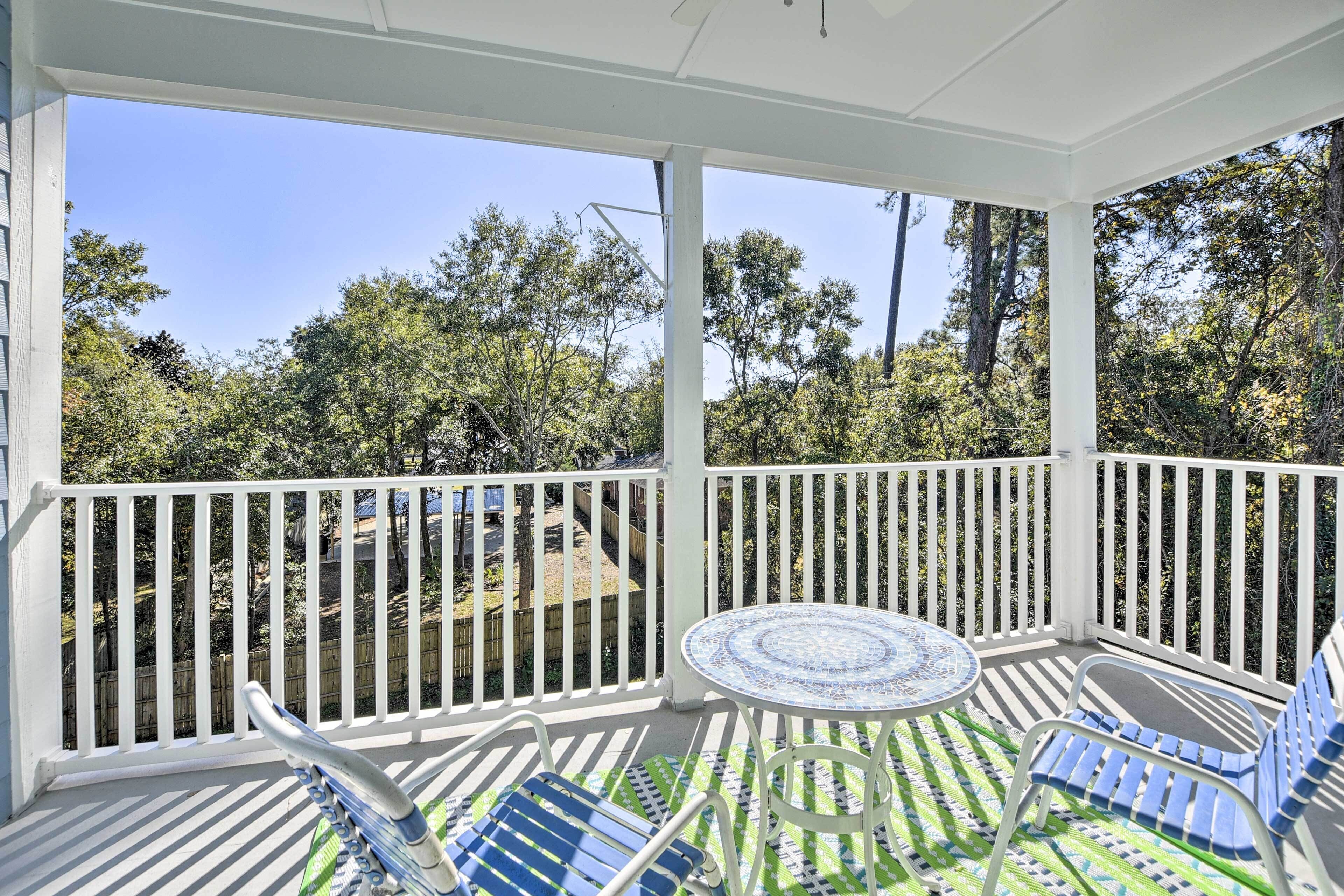 A covered outdoor patio with a round table and blue chairs, surrounded by white railings and a view of trees and a fence.