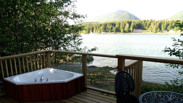 A wooden deck with a hot tub, a chair, and a table overlooking a lake with a forested mountain in the background.