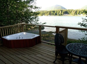 En träaltan med ett bubbelbad, en stol och ett bord, med utsikt över en sjö och ett skogbeklätt berg i bakgrunden.
