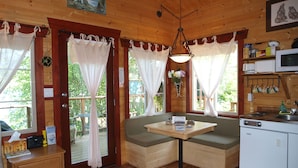 A cabin interior with a wooden dining table, bench seating, a kitchenette, and large windows with curtains.
