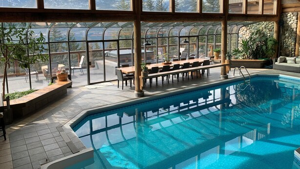 A pool area with a glass-enclosed terrace, wooden beams, and a view of snow-capped mountains. - New Denver Lodge – Relax, Unwind & Reconnect (New Denver)