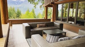 A modern outdoor seating area with a sofa and coffee table, wooden deck, and large windows offering mountain views. - New Denver Lodge – Relax, Unwind & Reconnect (New Denver)